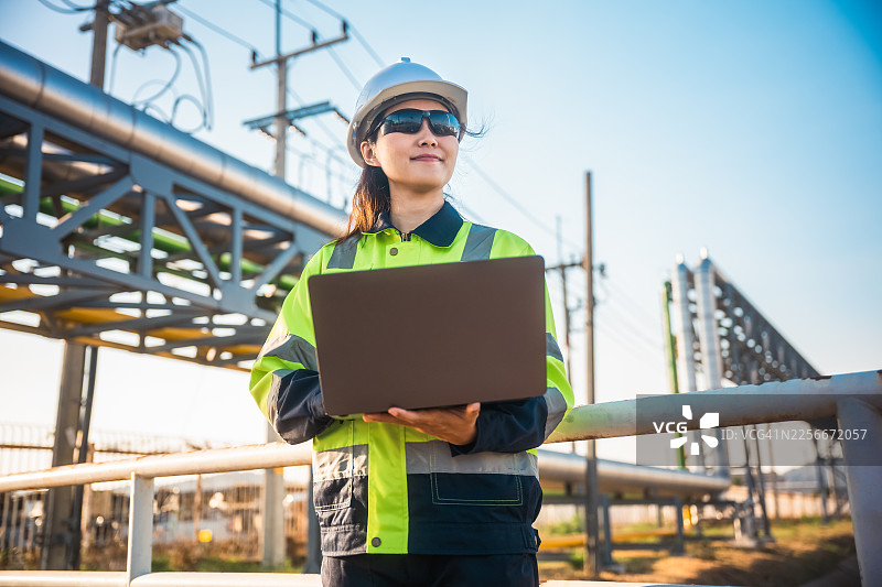 穿着安全帽的专业女性在工业设施中使用便携式电脑进行系统检查。石油炼油厂技术人员正在检查管道。图片素材