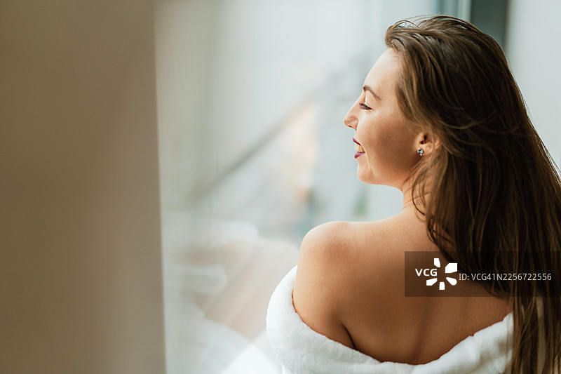 一位女子肩披毛巾，在柔和的自然光下放松地凝视窗外。图片素材