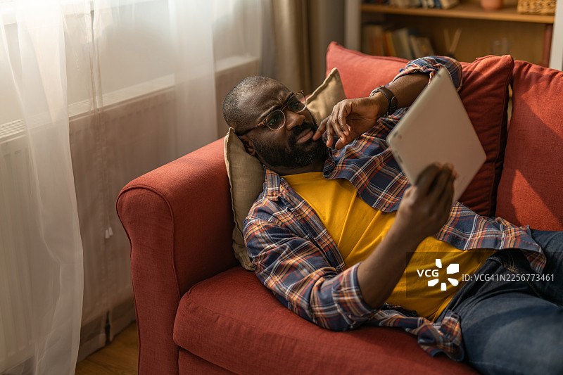 男人躺在沙发上，若有所思地使用平板电脑图片素材