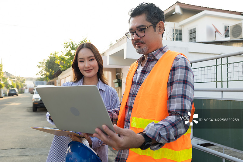 建筑经理和亚洲工程师在工地上用笔记本电脑监督建筑进展。身穿安全背心和头盔的亚裔男女工程师正在检查结构元素。图片素材