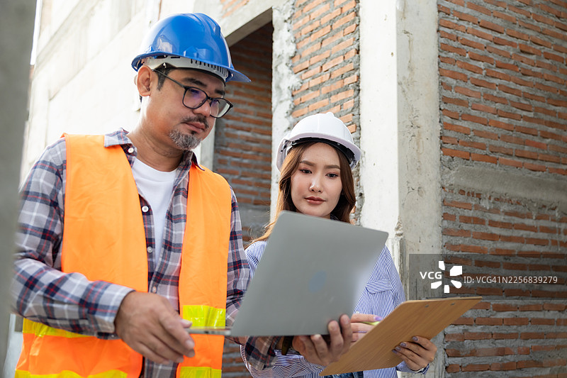 建筑经理和亚洲工程师在工地上用笔记本电脑监督建筑进展。亚洲男性和女性工程师穿着安全背心和头盔检查结构构件。图片素材