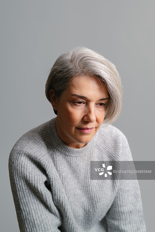 一位头发花白的成熟的女性，穿着灰色毛衣，若有所思地向下看，背景为中性色调。图片素材