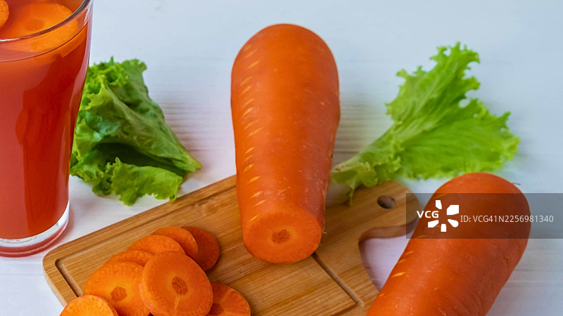 新鲜制作的胡萝卜汁，配有整根胡萝卜、胡萝卜片和生菜，摆放在白色桌子上，倡导健康饮食图片素材