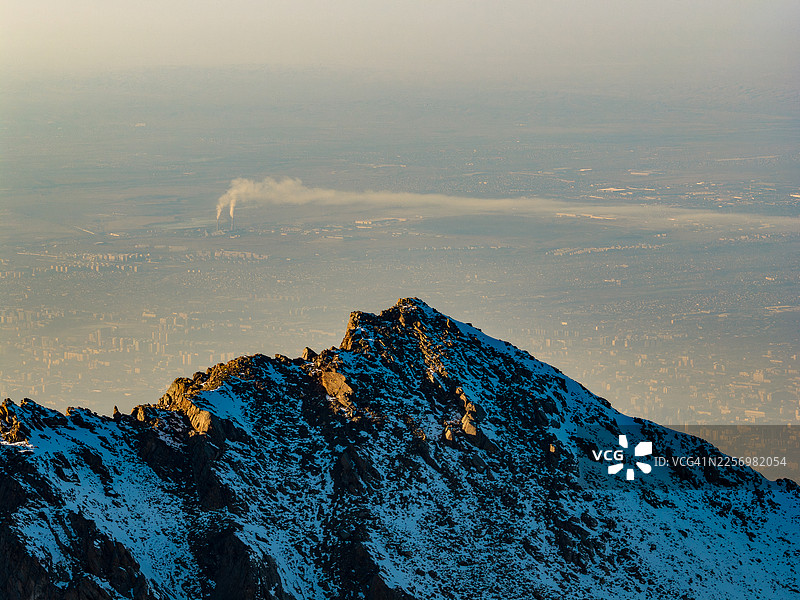 远处工业烟雾笼罩下的，宏伟的雪山之巅俯瞰着广阔而朦胧的城市景观，哈萨克斯坦阿拉木图，金堡拉克滑雪胜地图片素材