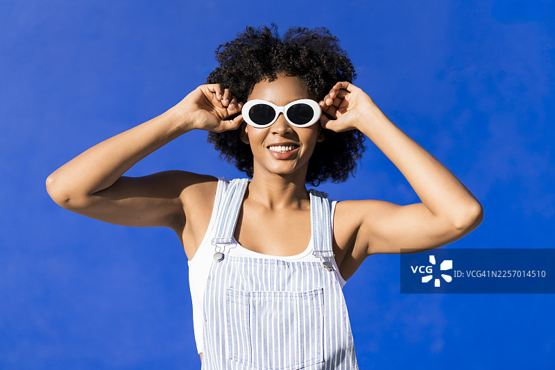 戴着太阳镜、留着卷曲非洲发型的快乐女子在蓝色墙壁前摆姿势图片素材