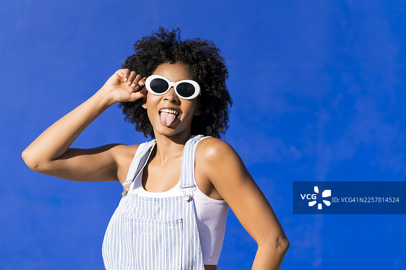 戴着太阳镜、留着非洲发型的俏皮女子在蓝色墙前快乐地摆姿势图片素材