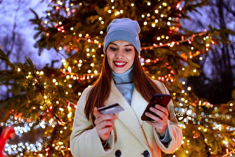 一位女士在圣诞树灯光下，手持手机和银行卡，面带微笑地进行网上购物图片素材