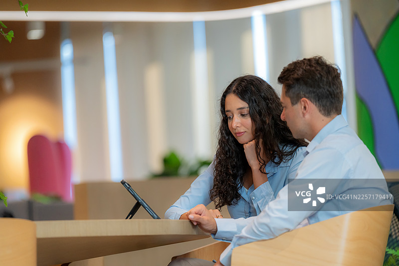 一位专业的男商人和一位女商人正在现代联合办公空间协作，并指向一台数字平板电脑。不同背景的办公室同事们在一个明亮的室内讨论创意项目想法和营销策略。图片素材