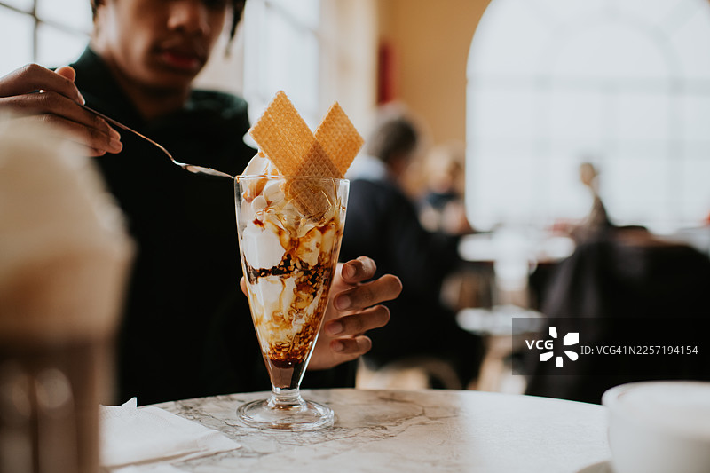一位年轻男子正在享用高脚杯中盛放的，含有坚果和焦糖风味的冰淇淋圣代。图片素材
