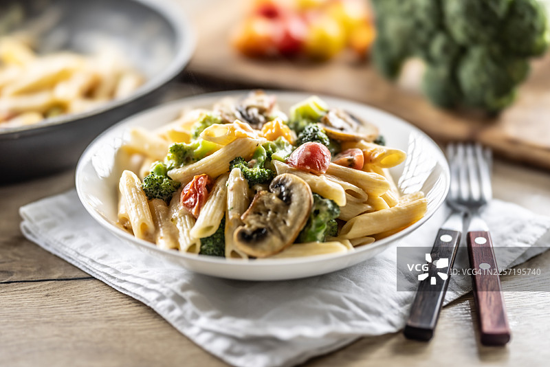 奶油酱笔管面特写，配有新鲜西兰花、蘑菇和圣女果，盛放在乡村风格的木桌上。生活方式摄影，背景柔和虚化，光线自然，非常适合美食和烹饪主题。图片素材