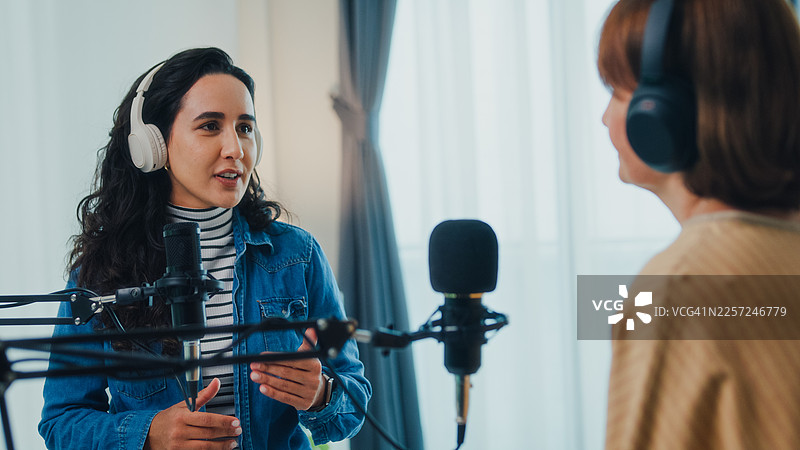 特写：成熟的白人女性和年轻的拉丁裔女性网红戴着耳机，在现代家庭工作室共同主持播客。直播播客节目和访谈概念。图片素材