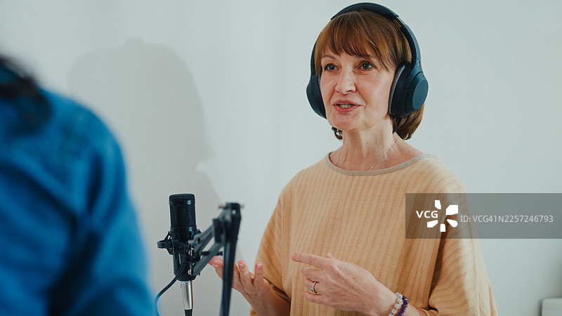 成熟的白人和年轻的拉丁裔女性网红戴着耳机，在现代家庭工作室共同主持播客。直播播客节目和访谈的概念。图片素材