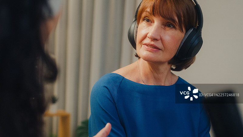 近距离拍摄，两位成熟的白人和年轻的拉丁裔女性网红戴着耳机，在夜晚的现代家庭工作室共同主持播客。直播播客节目和访谈概念。图片素材