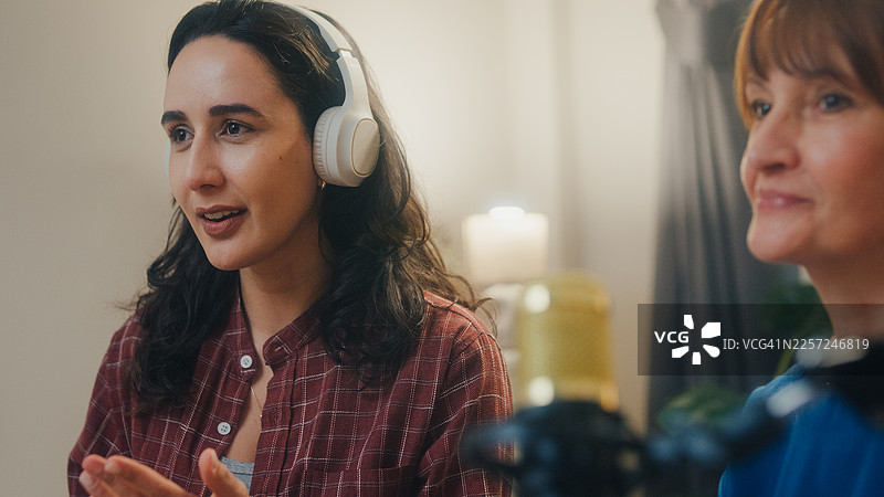 特写：年长的白人和年轻的拉丁裔女性网红戴着耳机，在夜晚的现代家庭录音室里一起主持播客。直播播客节目和访谈概念。图片素材