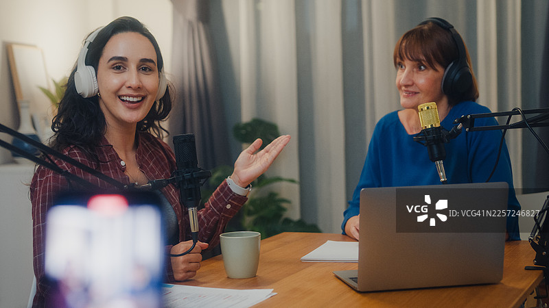 成熟的白人和年轻的拉丁裔女性网红戴着耳机，在夜晚现代化的家庭工作室直播播客节目。直播播客节目和访谈的概念。图片素材