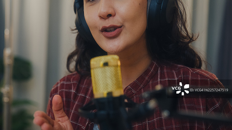 近景拍摄：年轻的拉丁裔女性网红戴着耳机，在夜晚的家庭工作室现场直播播客节目。直播播客节目和访谈的概念。图片素材