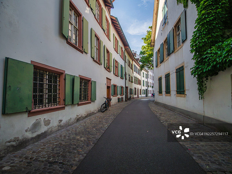 瑞士巴塞尔市中心老城区，一条铺设好的空旷步行街的广角景观，街道两旁是巴洛克风格的建筑和房屋，配有绿色百叶窗，有些还装饰着爬藤植物，路边停放着一辆自行车。图片素材