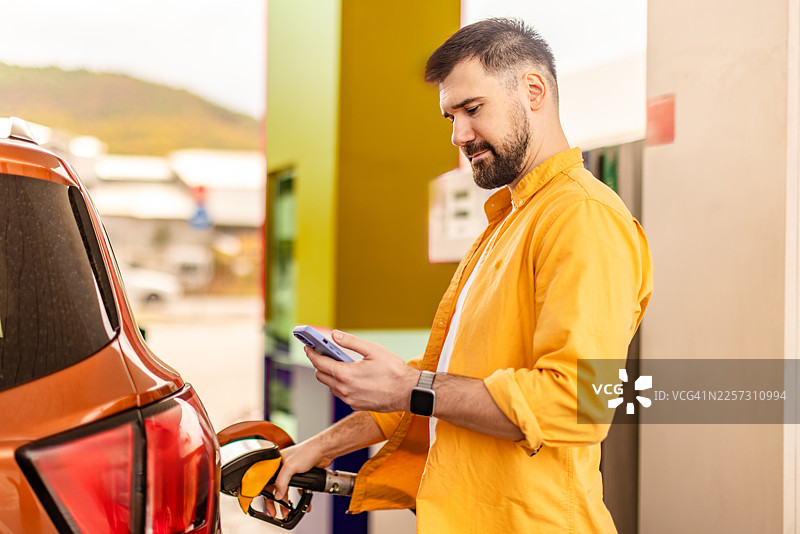 一名男子在繁忙的加油站为汽车加油，同时查看手机上的燃油价格和行程路线更新图片素材