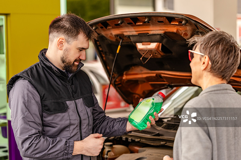 白天，一名男子在汽车加油站内向一名女子展示一瓶液体，同时处理加油需求。图片素材
