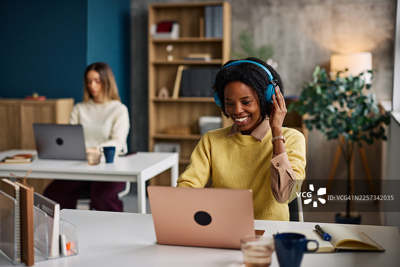 戴着蓝色耳机的快乐女子在现代协作式办公空间用笔记本电脑工作图片素材