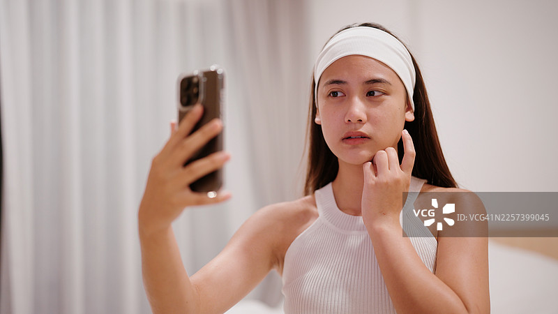 亚洲女性通过手机前置摄像头检查自己的皮肤，显得不太满意图片素材