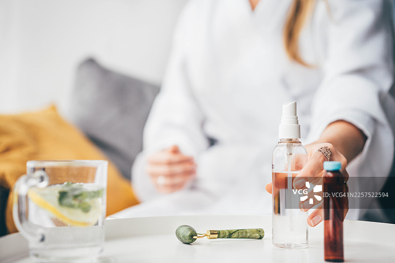 一位身穿白色浴袍的女士正伸手去拿托盘上的护肤品瓶子，托盘上还放着玉石滚轮和柠檬水。图片素材