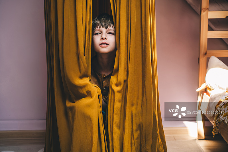 一个穿着黄色衣服的小男孩，藏在一个米黄色的布秋千里，这个场景在一个温馨的儿童房里。柔和的光线和暖色调营造出一种宁静舒适的氛围。图片素材