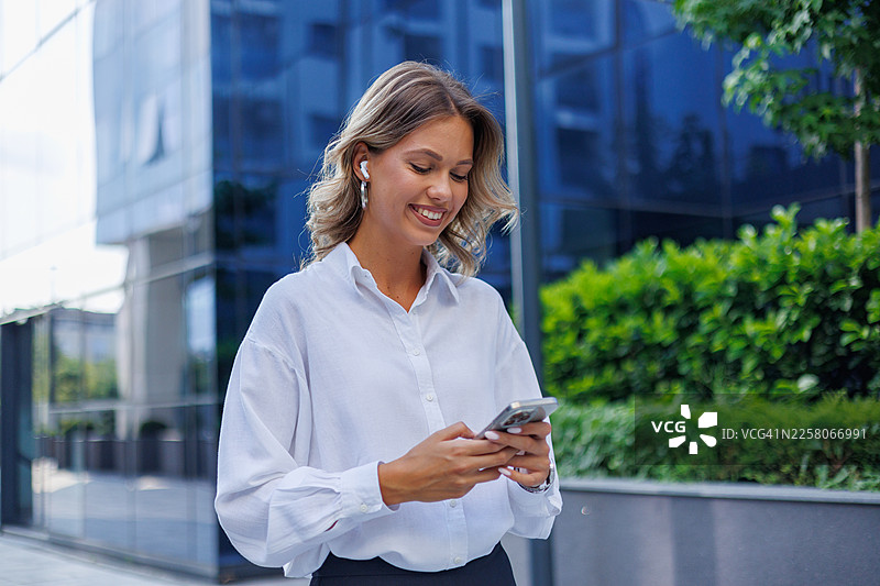 年轻的女商人微笑着走在现代办公楼外，使用手机和无线耳机图片素材