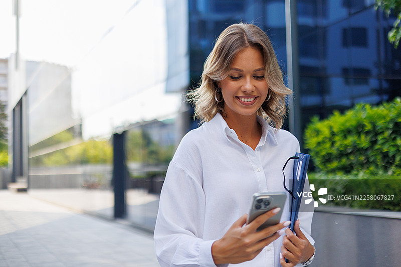 一位女商人在户外微笑着看着智能手机，在城市环境中进行连接和交流图片素材