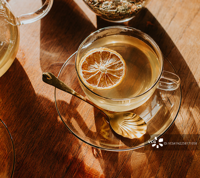 阳光下，木桌上的玻璃杯中盛着草本茶，旁边放着一片干柑橘片。图片素材
