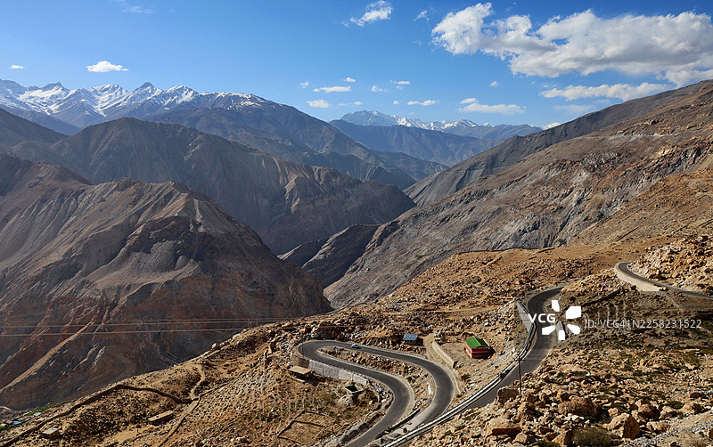 蜿蜒的山路，在一片崎岖干旱的土地上，坐落着一座小建筑，天空湛蓝，远方是白雪皑皑的山峰，印度喀扎，斯皮提谷图片素材