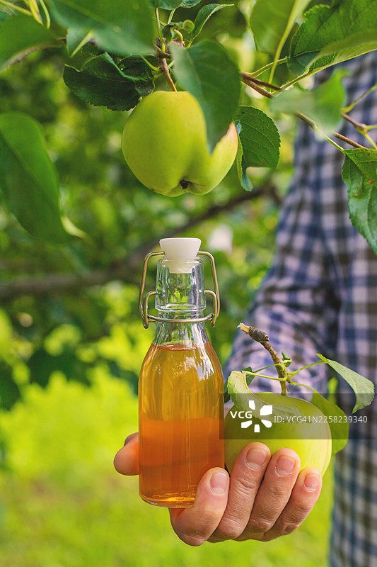 男人在果园的苹果树下，手里拿着一瓶新鲜的苹果汁和一颗青苹果图片素材