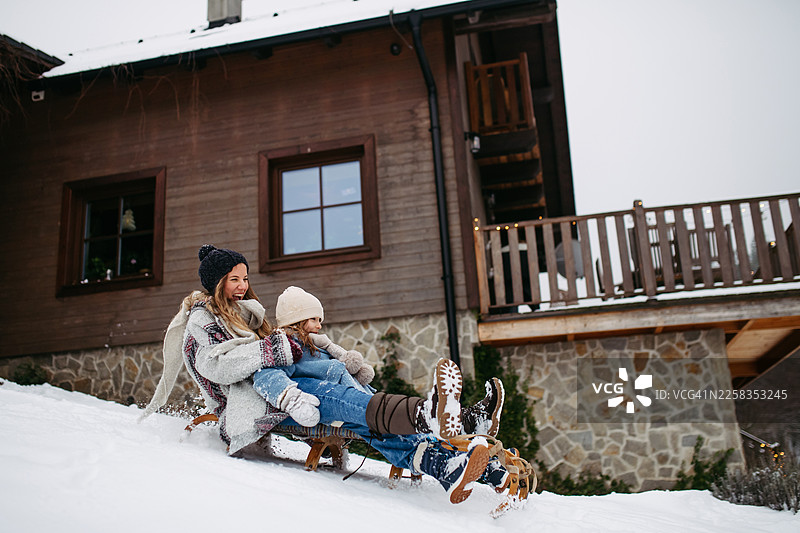 女孩和妈妈在房子旁边的雪山上滑雪橇。图片素材