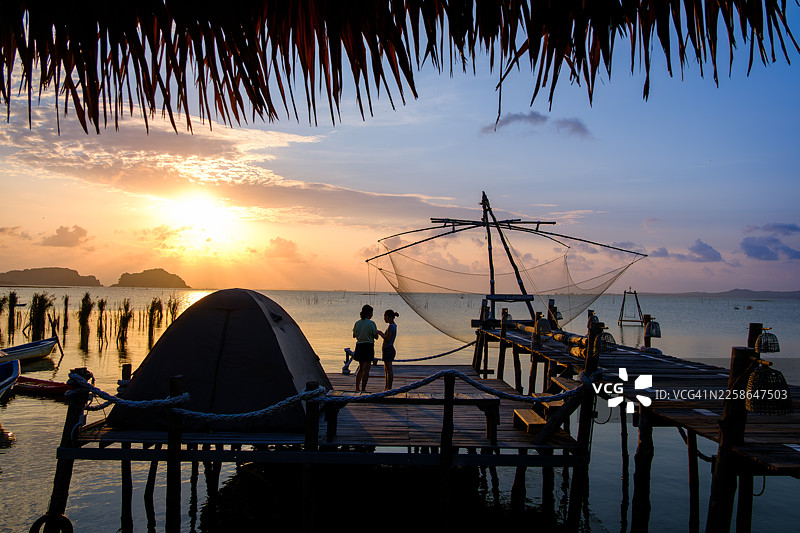 日落时分，女子坐在木码头上，两侧停靠着两艘皮划艇，俯瞰后景，地点为泰国宋卡湖、薄他仑府玛岛。图片素材
