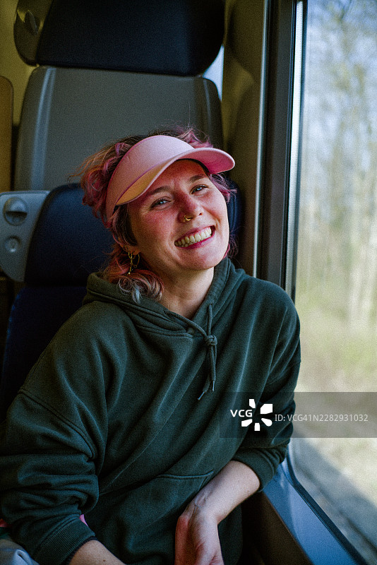 白天旅行时，戴着粉色遮阳帽和连帽衫的微笑女子坐在火车窗户旁图片素材