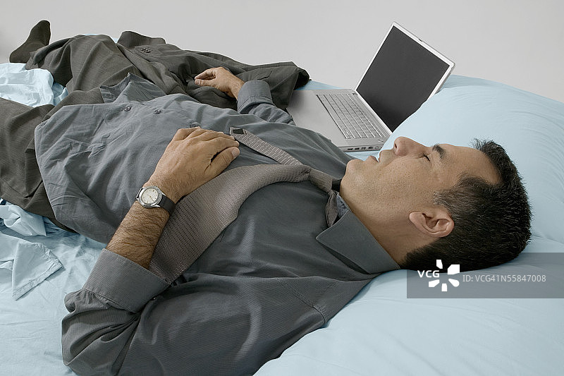 疲惫的商人穿着整齐地在床上睡觉，旁边放着笔记本电脑图片素材