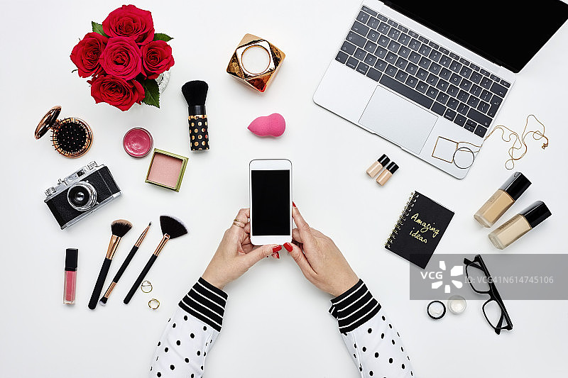 博主手持智能手机，周围摆放着美妆产品图片素材
