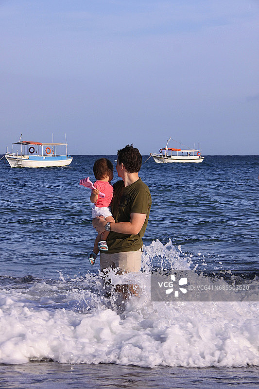 父亲抱着蹒跚学步的女儿在海滩边玩耍图片素材