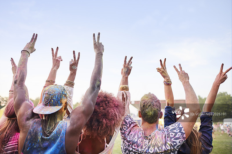 一群朋友在节日上，身上沾满彩色粉末，举起手臂，用手指做出和平标志（后视图）。图片素材