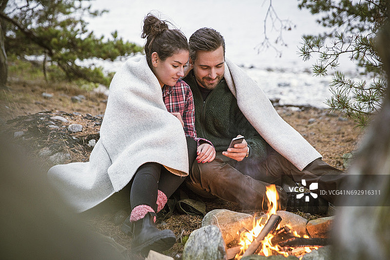 情侣在露营地篝火旁裹着毯子使用手机图片素材