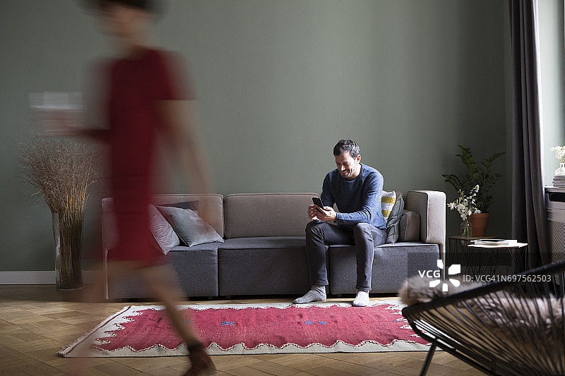 男人坐在客厅沙发上，女友从前景经过图片素材