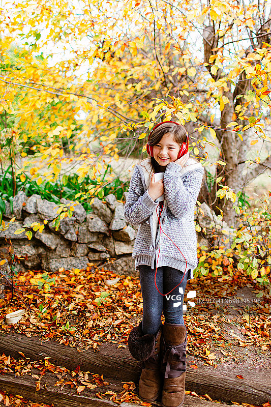 秋天，在色彩鲜艳的树下戴着耳机听音乐的年轻女孩图片素材