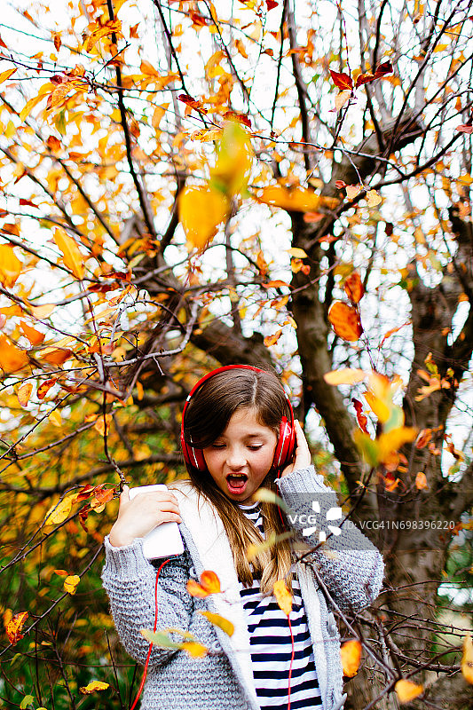 秋天，在色彩鲜艳的树下戴着耳机听音乐的年轻女孩图片素材