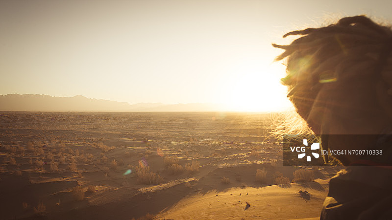 在伊朗中部梅斯尔附近的沙漠中观看日落的，有脏辫的年轻冒险家图片素材