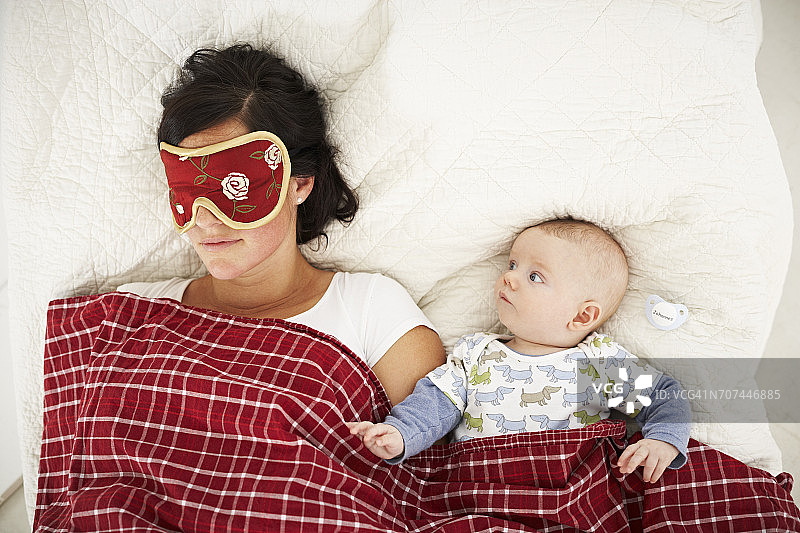 戴着睡眠面膜的母亲和醒着的婴儿躺在床上图片素材