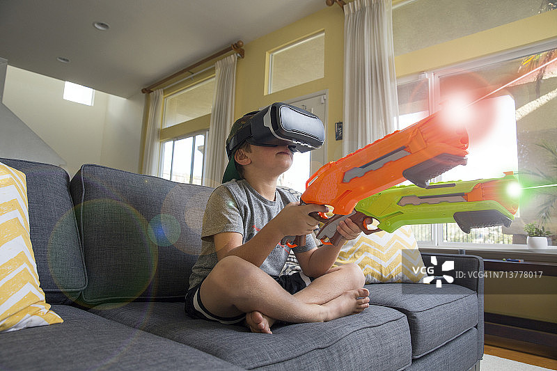 盘腿坐在沙发上，戴着VR头显，拿着激光枪的男孩图片素材