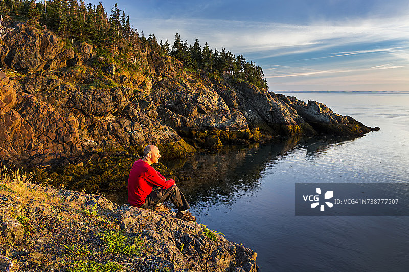 男人坐在日出时多岩石的海岸线上，有蓝天和白云图片素材
