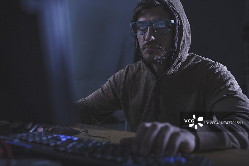 表情严肃的窃贼穿着连帽衫在桌旁使用电脑图片素材