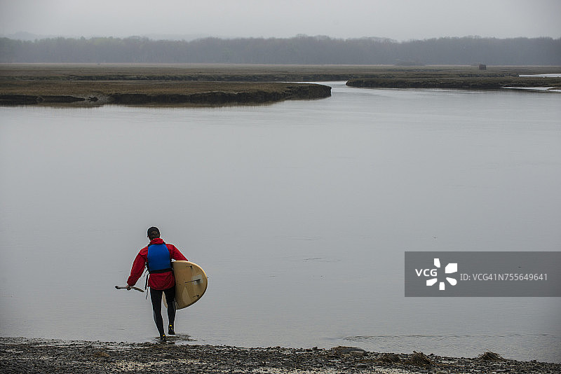 男人手持立式桨板走向湖边，地点：美国缅因州斯卡伯勒沼泽图片素材