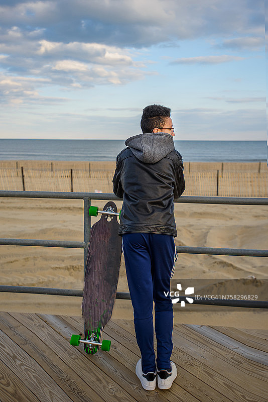 倚靠在栏杆上，看着风景的男孩，滑板在他旁边，美国新泽西州阿斯伯里图片素材